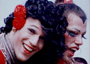 Still from the film Gay USA. A close up of two people smiling at a pride march in the 70's. They are both wearing vivid red lipstick. One person wears red flowers in their log black curly hair. The other person has big fluttery false eyelashes and a painted hairline.